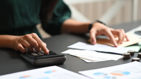 A woman is managing expenses and receipts using a calculator while sitting at a desk. - JacRox